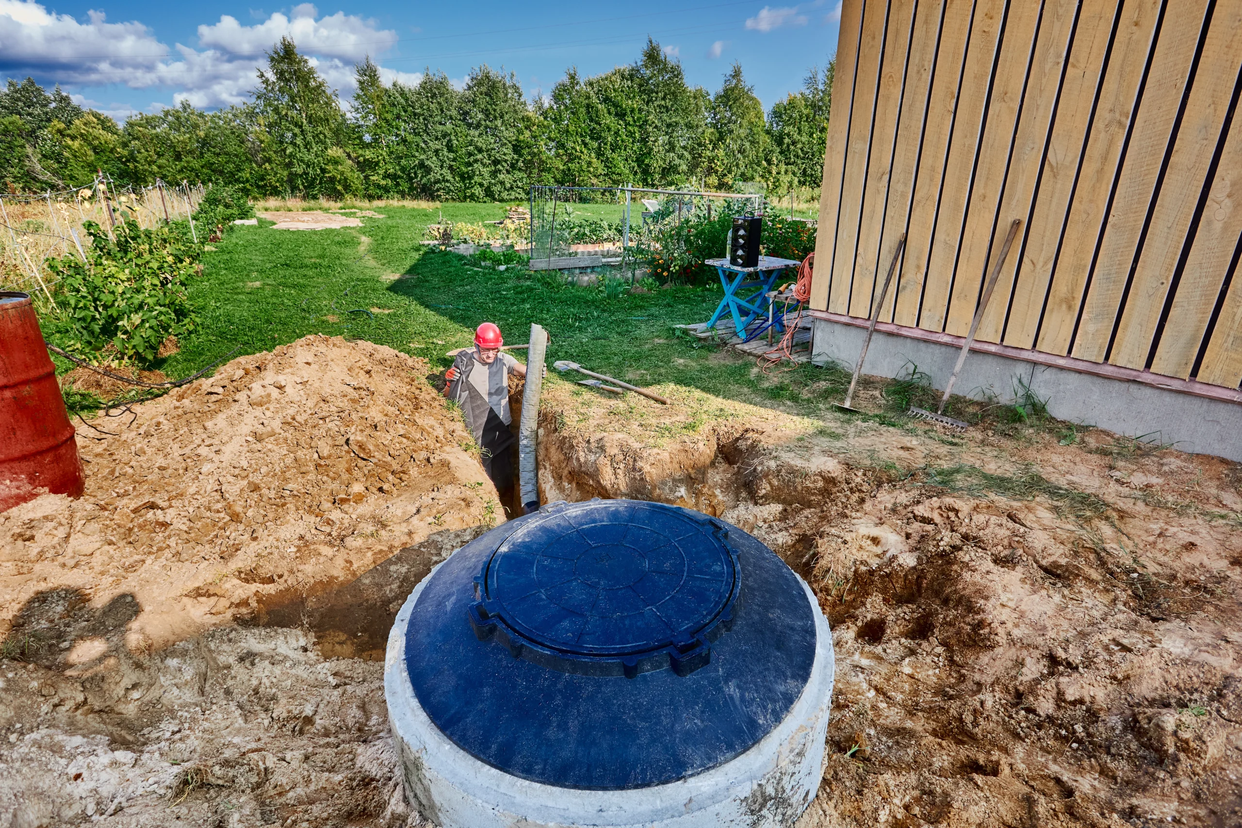 Worker installing a bio septic tank in a residential backyard, with the tank partially buried in the ground near a wooden house and surrounded by construction tools and soil. Crystal Bio Septic Tank