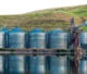 Row of large industrial metal silos with blue conical roofs, used for storage, situated near a hillside and reflected in the calm water in front. Crystal Bio Septic Tank