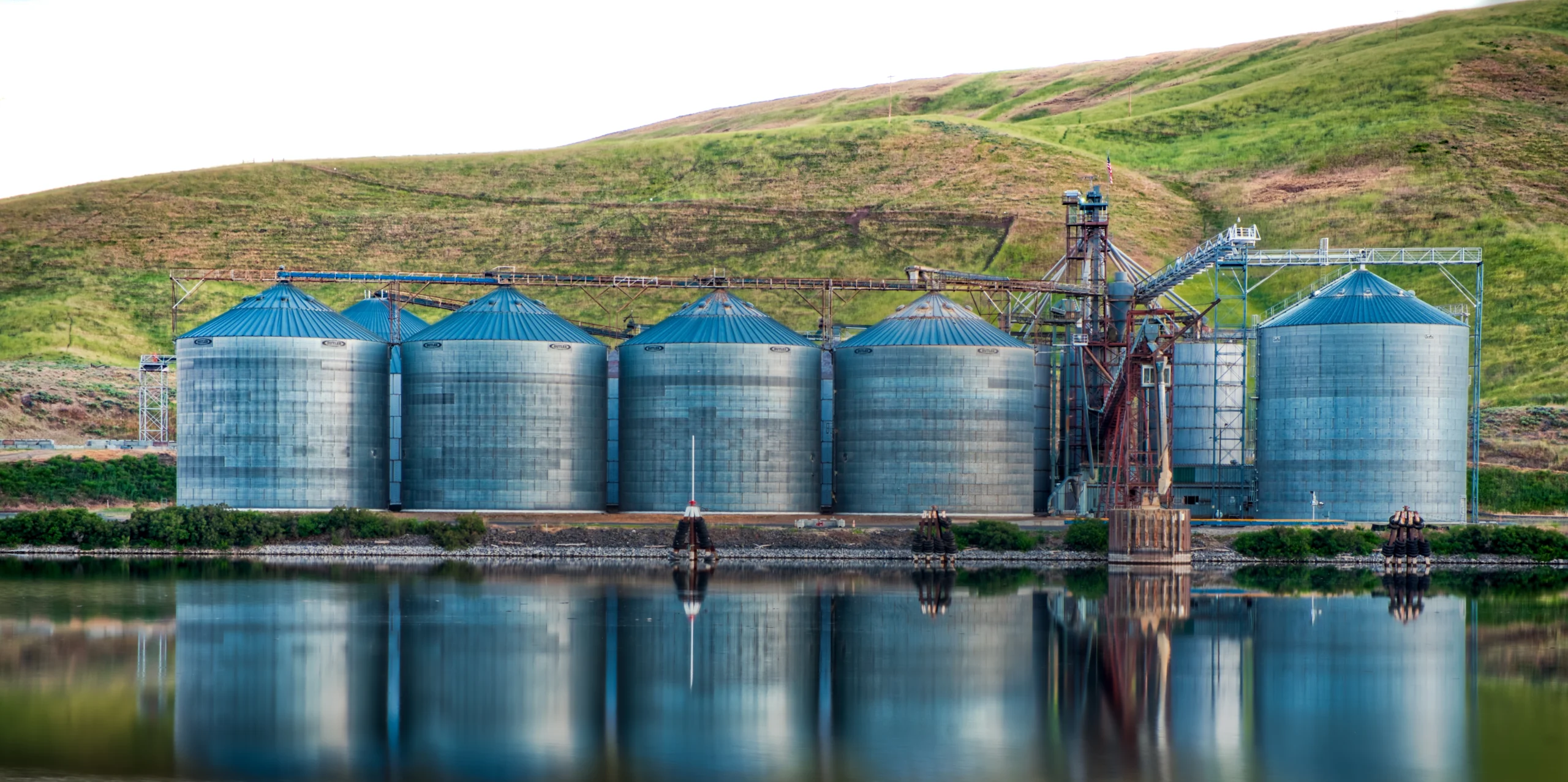 Row of large industrial metal silos with blue conical roofs, used for storage, situated near a hillside and reflected in the calm water in front. Crystal Bio Septic Tank